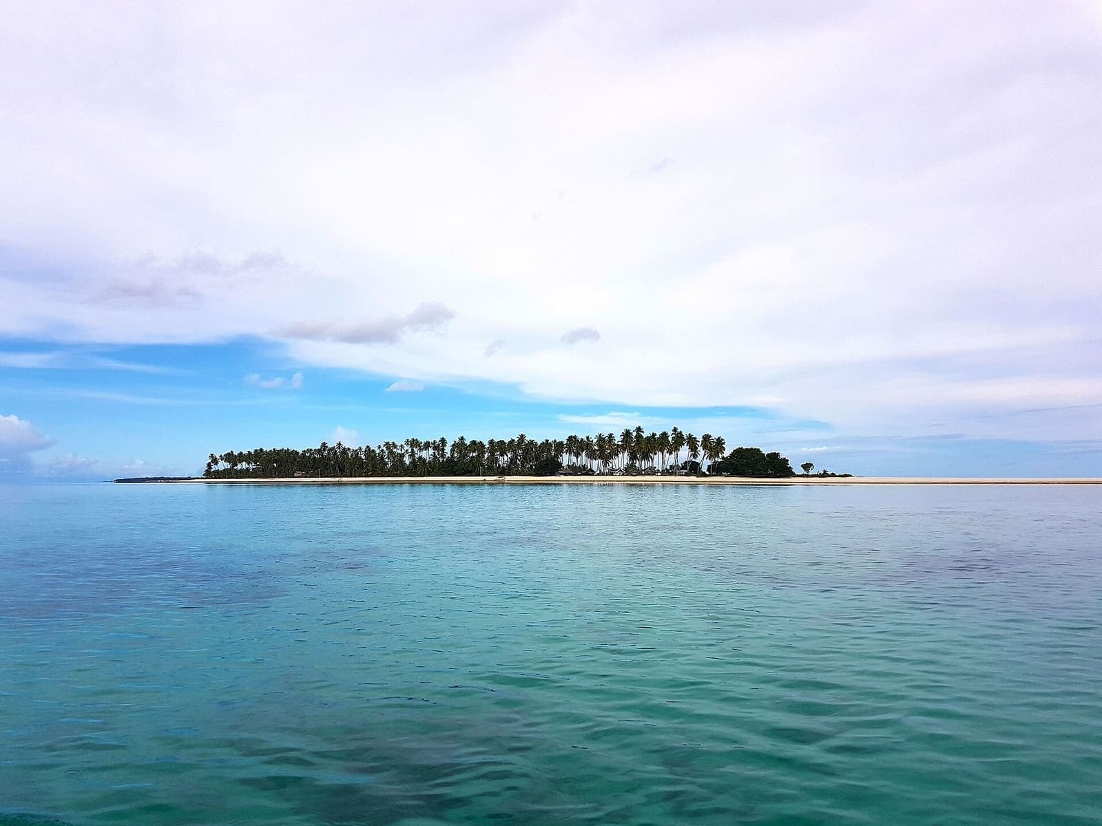 A low green island ringed by a pale sandbar in turquoise water under a wide sky.